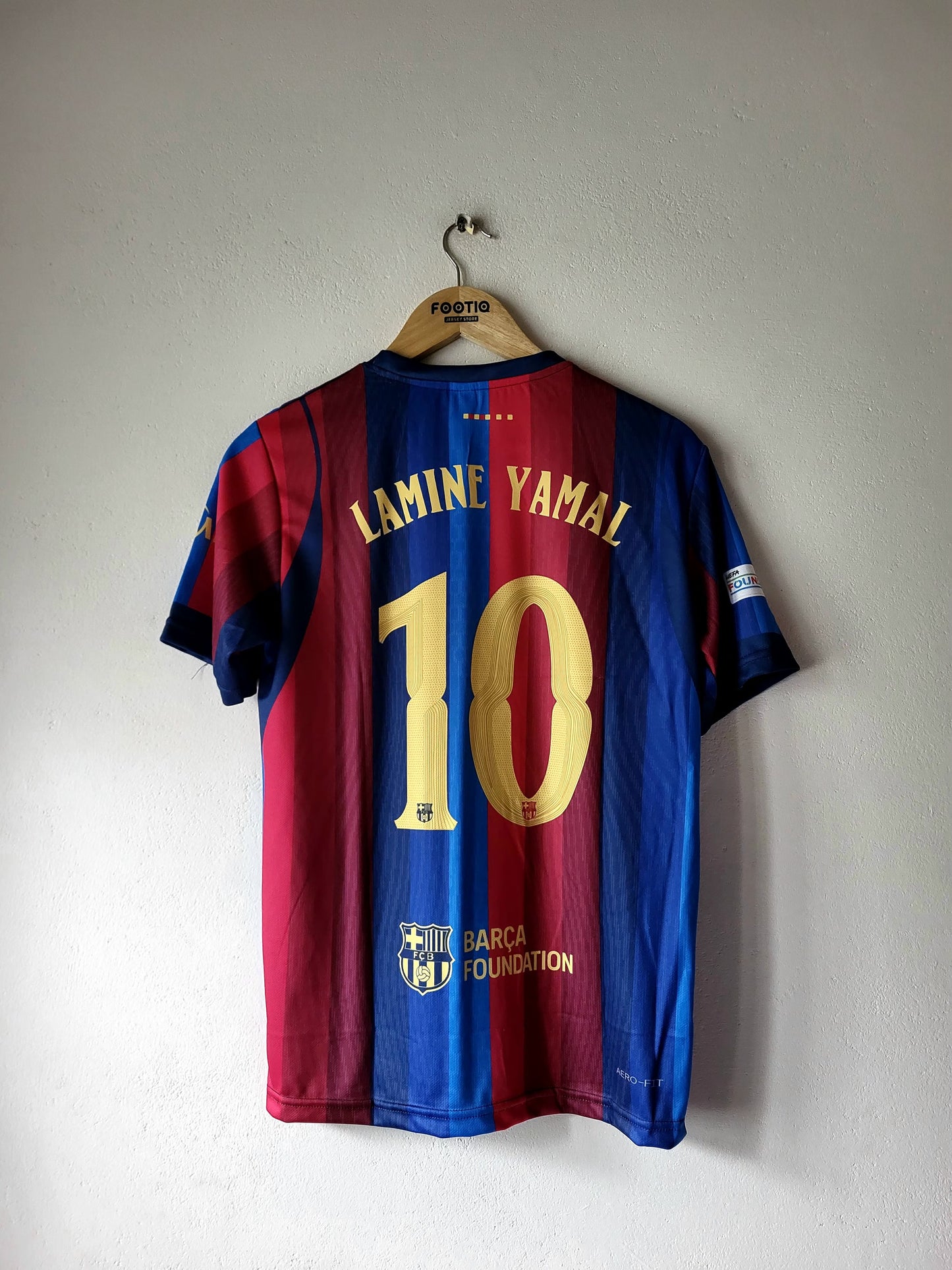 A Barcelona soccer jersey with the name Lamine Yamal and number 10 on the back, hanging on a wooden hanger against a plain wall.
