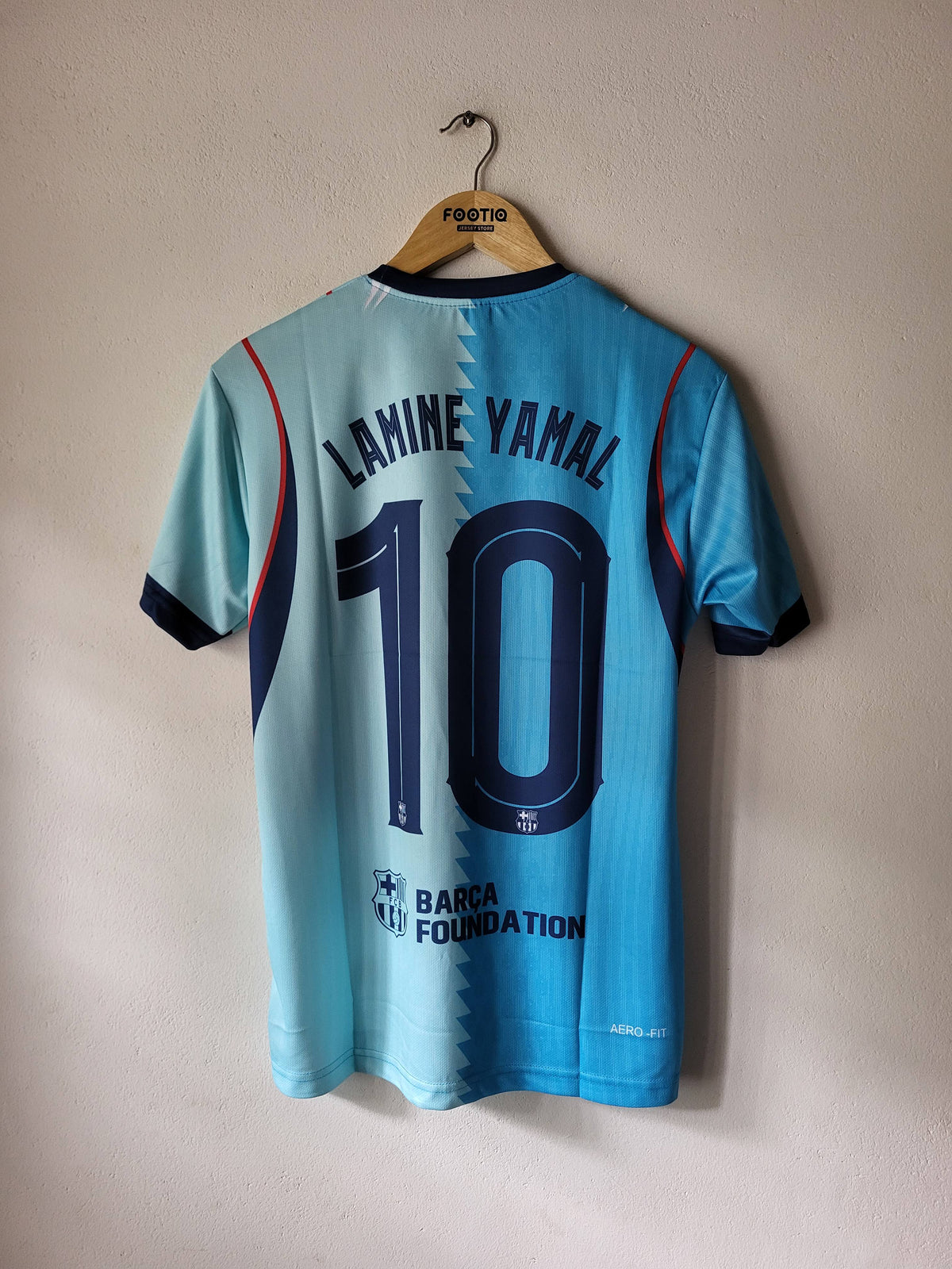 A light blue FC Barcelona soccer jersey with the name 'LAMINE YAMAL' and number '10' printed on the back, hanging on a wooden hanger against a textured beige wall.
