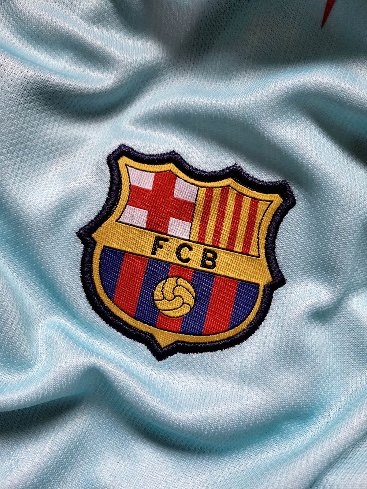 A close-up of the embroidered FC Barcelona crest patch on a light blue athletic jersey fabric.