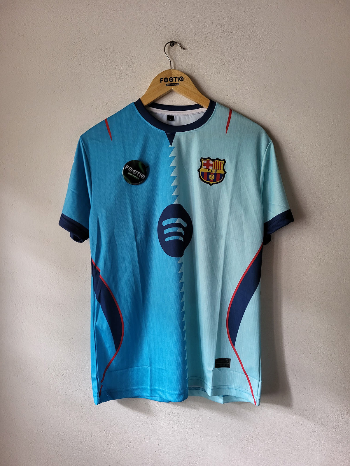 A light blue FC Barcelona soccer jersey hanging on a wooden hanger against a textured beige wall. The jersey features the club's crest on the left chest, a circular dark blue logo in the center, and dark blue sleeve cuffs and side panels with thin red piping details.