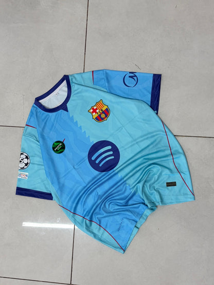 A light blue FC Barcelona soccer jersey with dark blue trim, laid flat on a light gray tiled floor. The jersey features the club's crest on the chest, a large Spotify sponsor logo, and a Champions League patch on the sleeve.