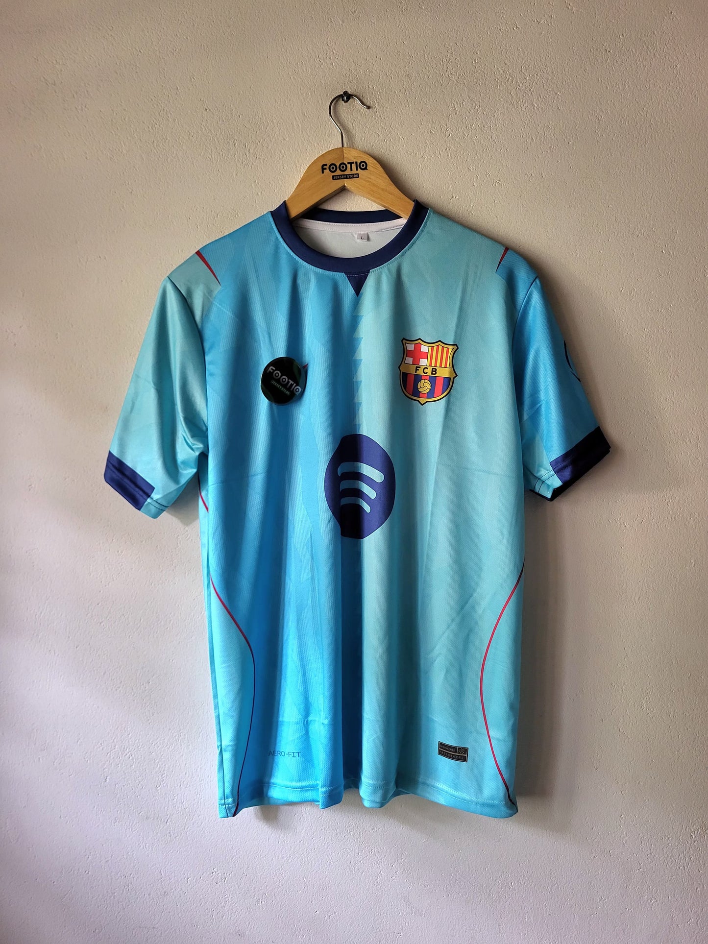 A light blue FC Barcelona soccer jersey hanging on a wooden hanger against a textured beige wall. The jersey features the club's crest on the left chest, a dark blue circular patch on the right chest, and a large dark blue Spotify logo on the center front.