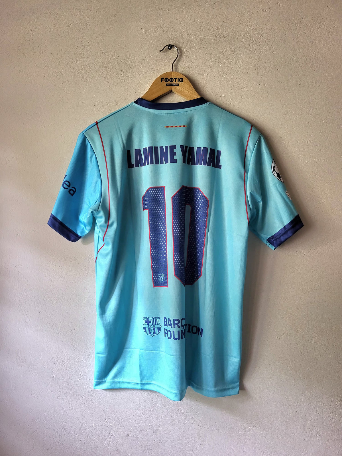 A light blue FC Barcelona soccer jersey with 'LAMINE YAMAL' and the number '10' printed on the back, hanging on a wooden hanger against a beige wall.