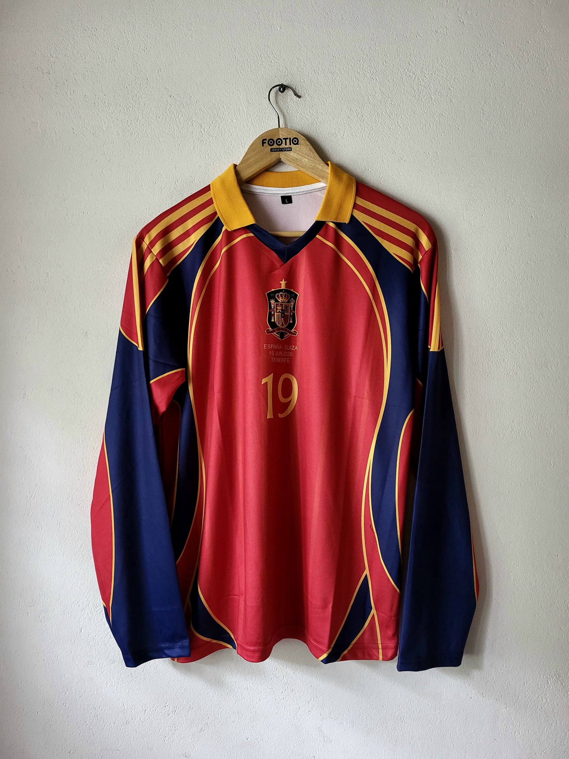 A long-sleeve Spain soccer jersey with red, navy blue, and yellow colors, hanging on a wooden hanger against a plain wall. The jersey features the Spanish national team crest and the number 19 on the front.