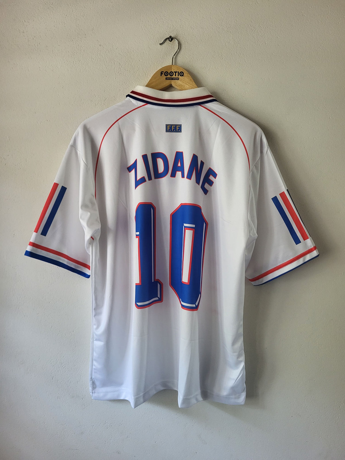 A white France national football team jersey with 'ZIDANE' and the number '10' printed on the back, hanging on a wooden hanger against a plain wall.