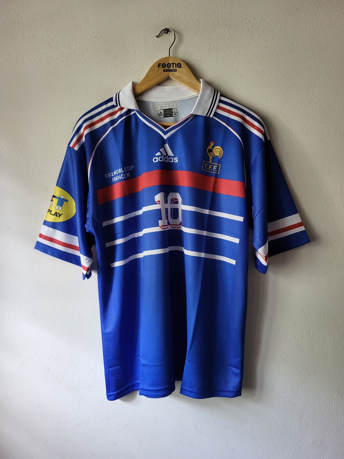 A blue France national football team Adidas jersey with red and white stripes, hanging on a wooden hanger against a plain wall. The jersey features the number 10 on the front, the Adidas logo, and the FIFA crest.