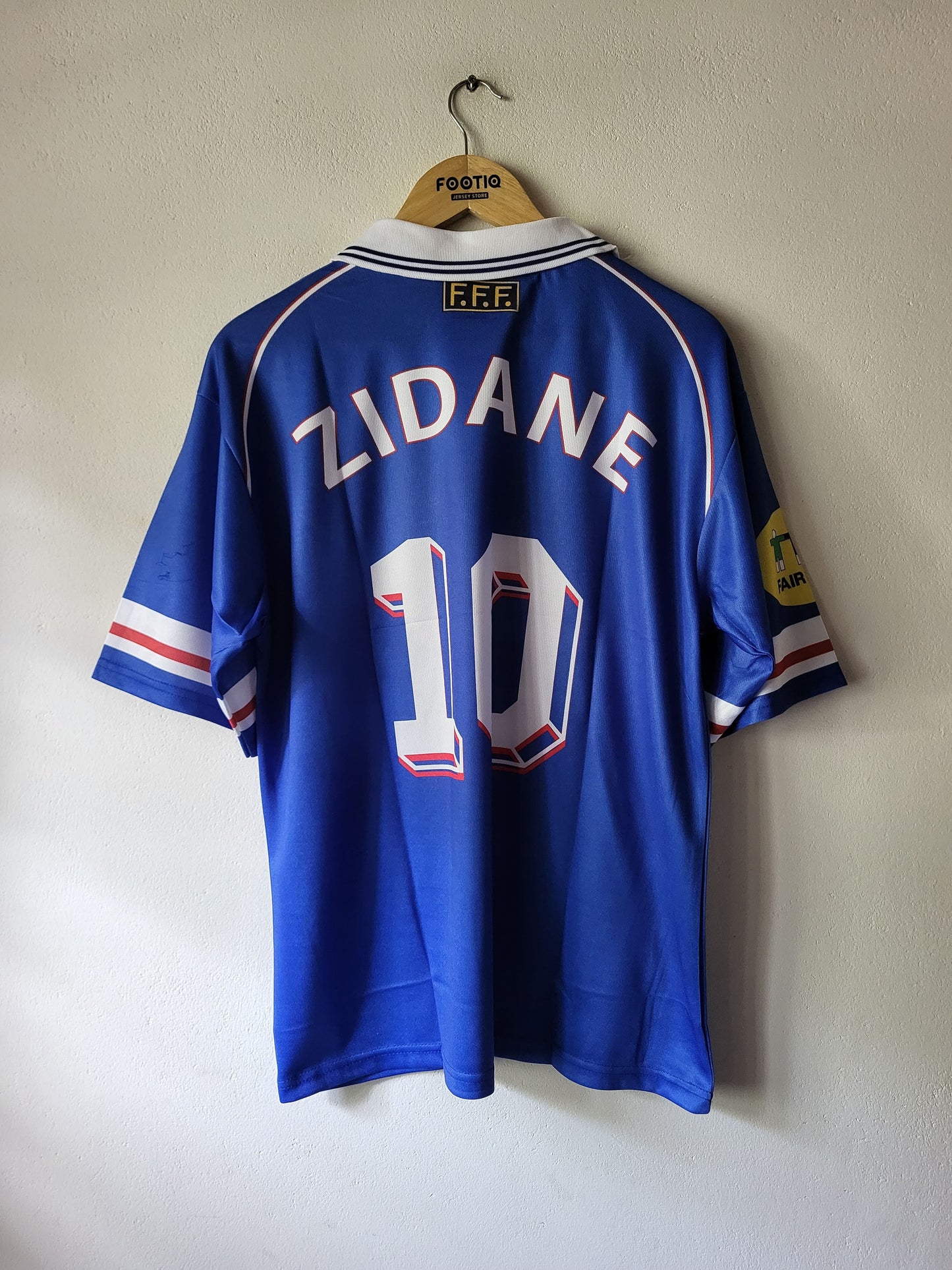 A blue France national football team jersey with 'ZIDANE' and the number '10' on the back, hanging on a wooden hanger against a plain wall.