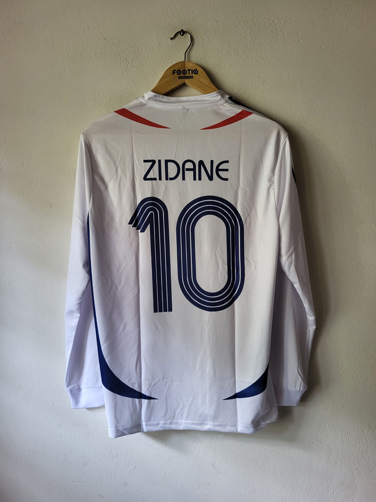 A white long-sleeve soccer jersey hanging on a wooden hanger against a plain beige wall. The back of the jersey features the name 'ZIDANE' and the number '10' in dark blue, with red accents near the shoulders and blue side graphics.