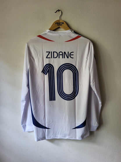 A white long-sleeve soccer jersey hanging on a wooden hanger against a plain beige wall. The back of the jersey features the name 'ZIDANE' and the number '10' in dark blue, with red accents near the shoulders and blue side graphics.